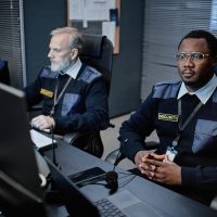 Three middle aged security guards including Black man and two Caucasian men monitoring surveillance screens in control room, sitting at desks with focused expressions, wearing uniforms and badges