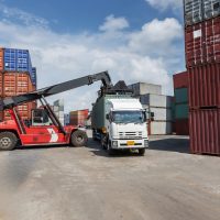 A red container forklift loads a cargo container onto a white truck in a busy shipping yard. Surrounded by stacked containers, the scene represents global logistics, trade, and freight transportation.