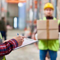 Portrait of two shipping workers marking shipping list and carrying cardboard box for delivery in modern warehouse